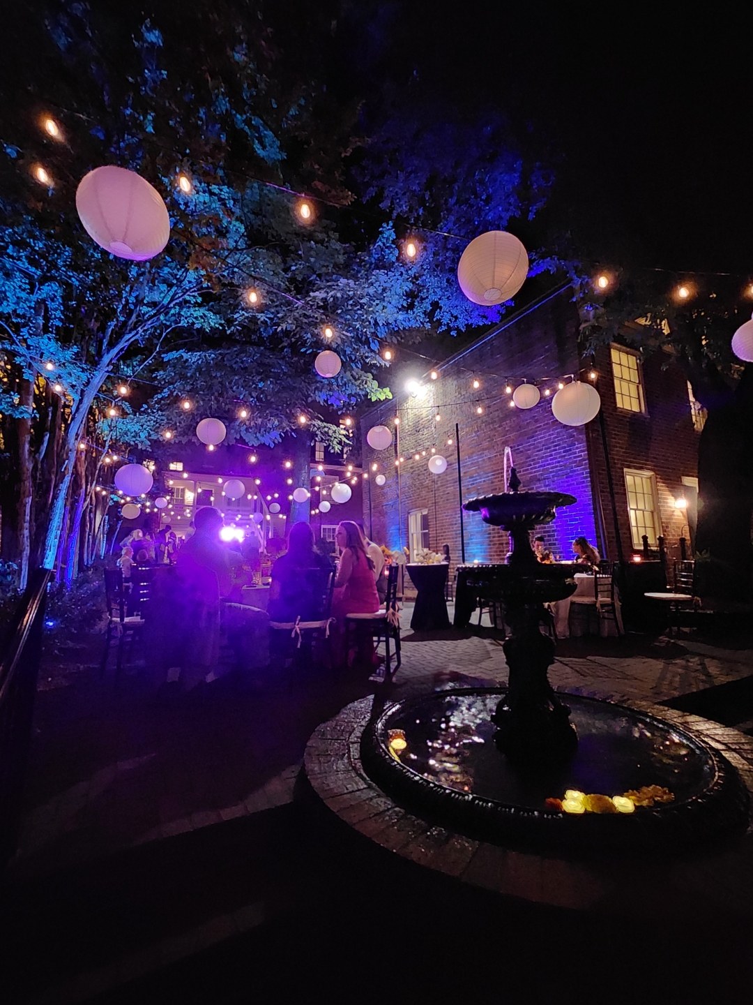 Courtyard reception at night lit with paper lanterns and string lights above a stone fountain