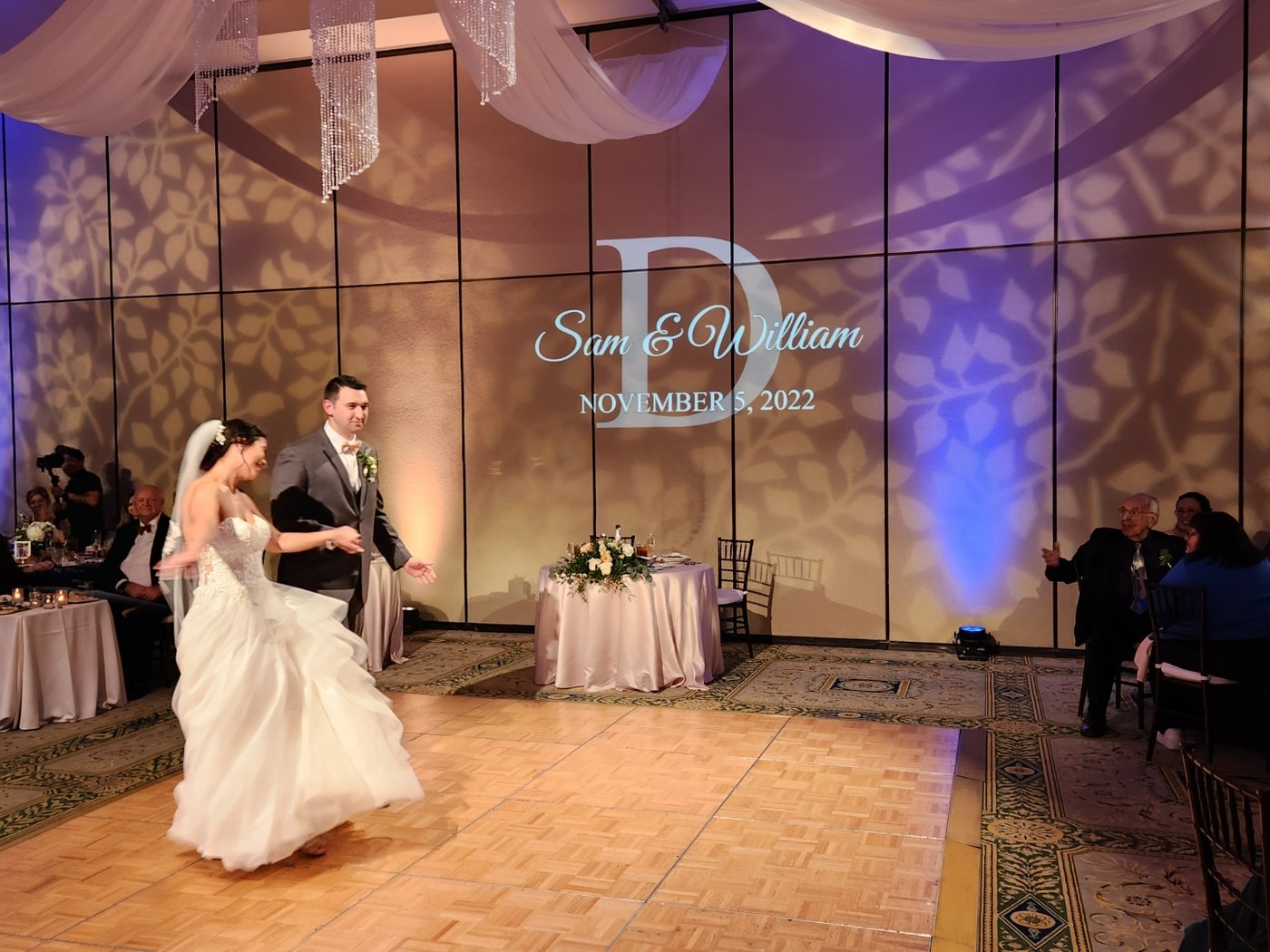 Couple entering a ballroom under a projected monogram with amber and lavender uplighting